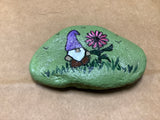 Gnome with pink flower in grass with background green rock. Made by local artist Cecelia. Each rock is $3.